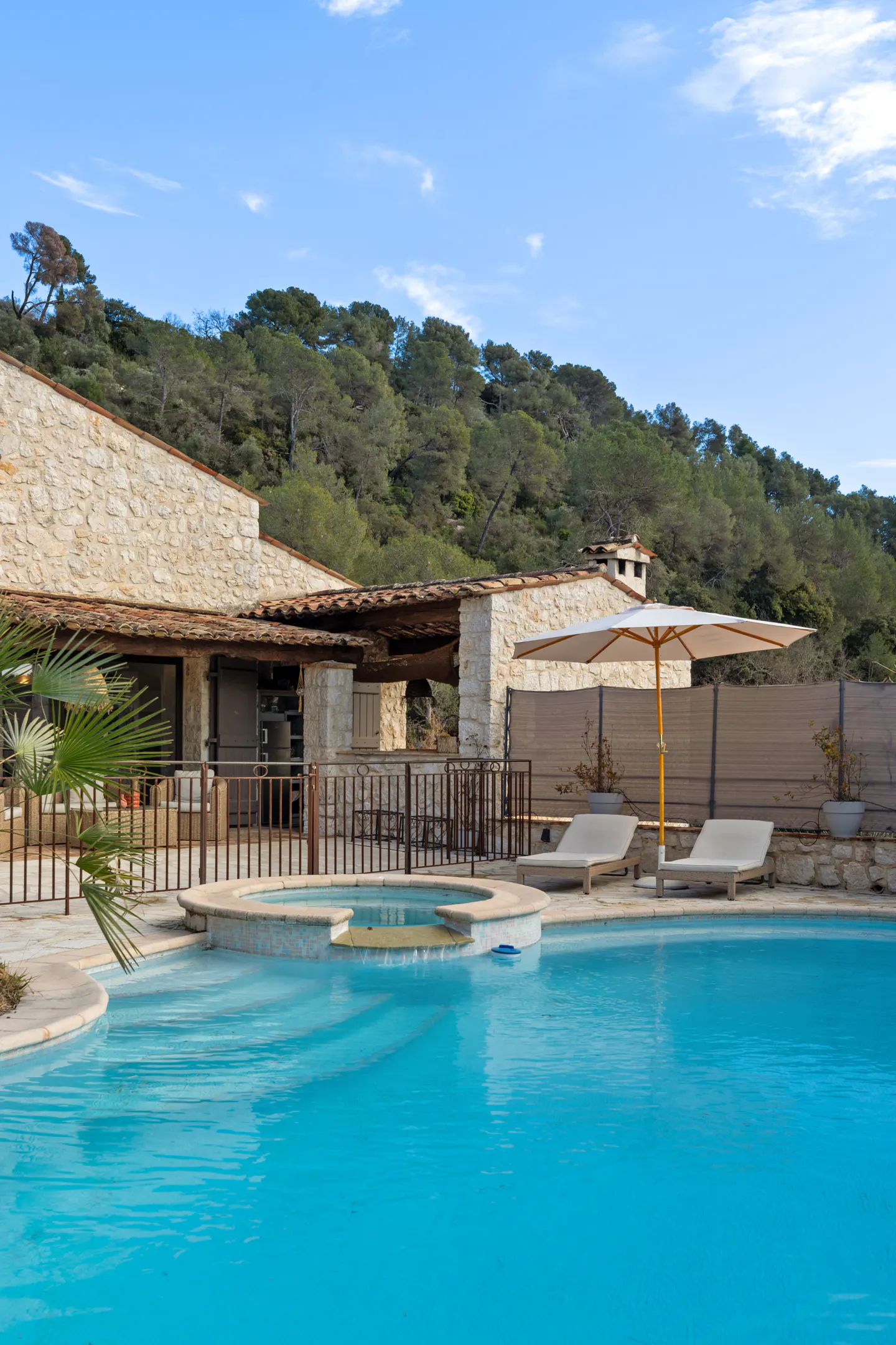 A turquoise pool with lounge chairs and an umbrella in front of a stone house and green trees.