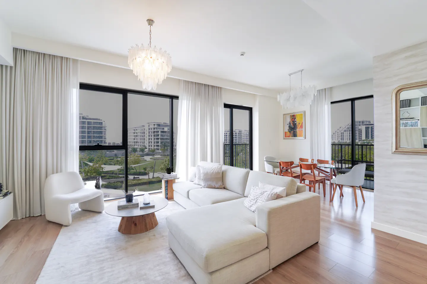 Bright, modern living room with white sectional sofa, wood floors, and large windows overlooking a park and buildings. Chandelier and dining set visible.