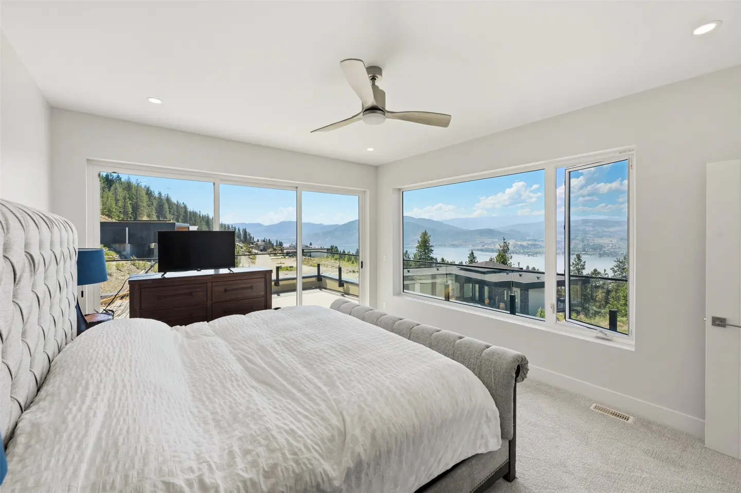 Modern bedroom with a grey tufted bed, dark wood dresser, and large windows showcasing a scenic lake and mountain view.