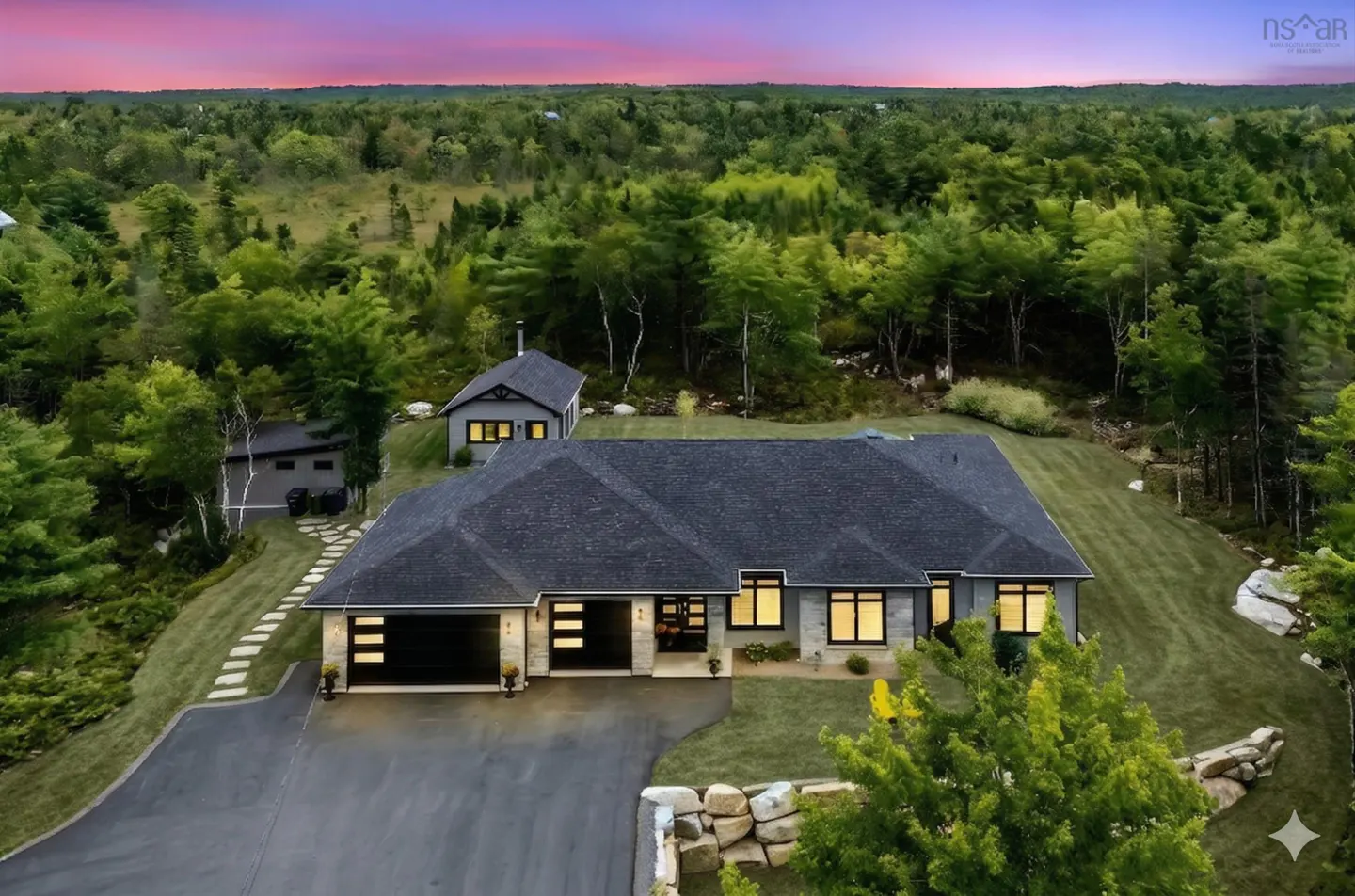 Aerial view of a gray stone house with a black roof, surrounded by green trees and a pink and purple sky.