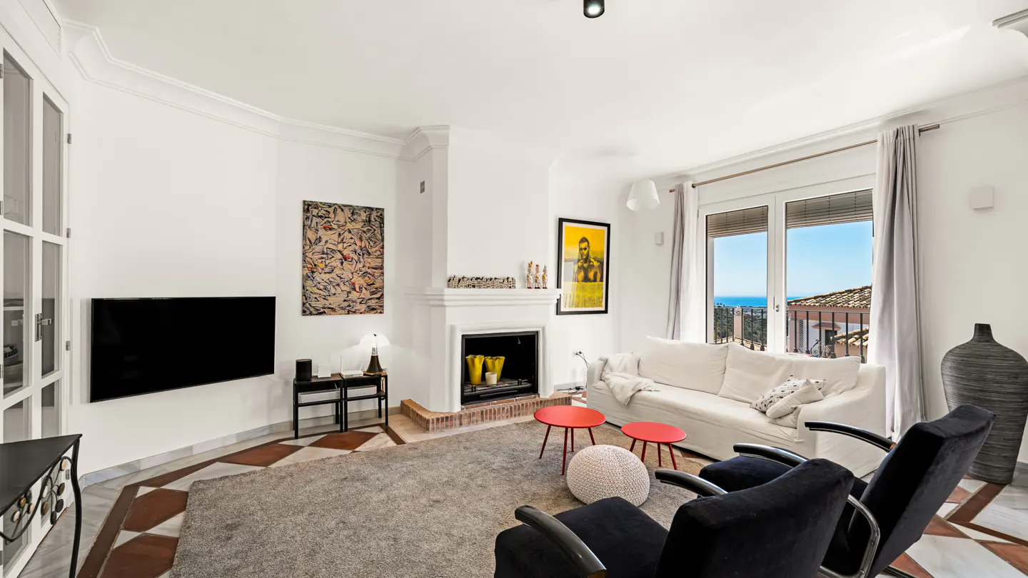 Bright living room with white walls, fireplace, TV, and art. Two black chairs face a white sofa near a window with an ocean view.