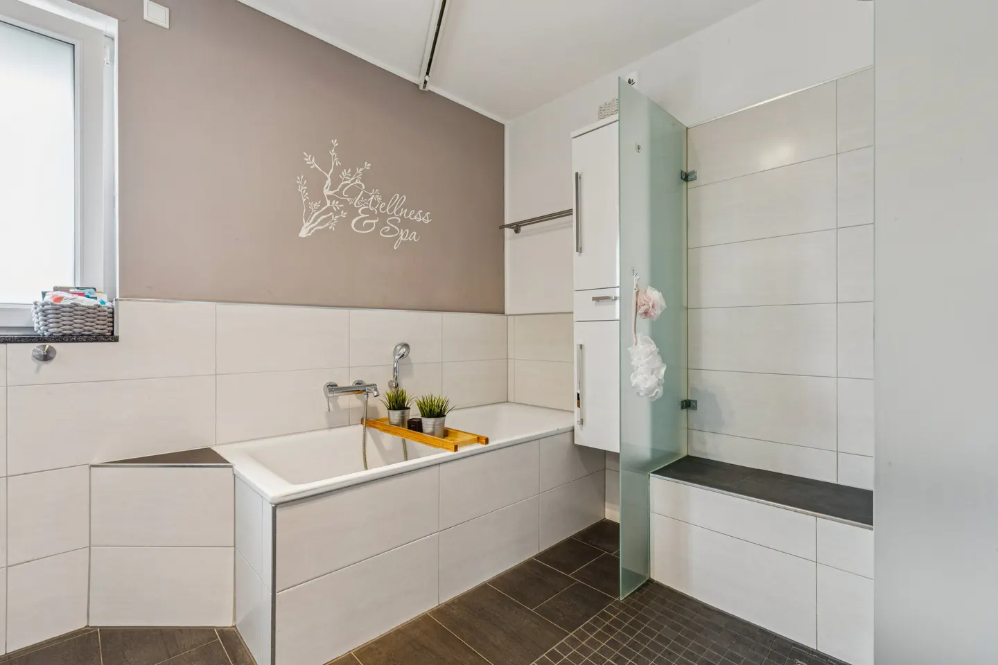 A modern bathroom with white tiles, a bathtub with plants, and a glass-enclosed shower with a bench. The wall is taupe with "Wellness & Spa" text.