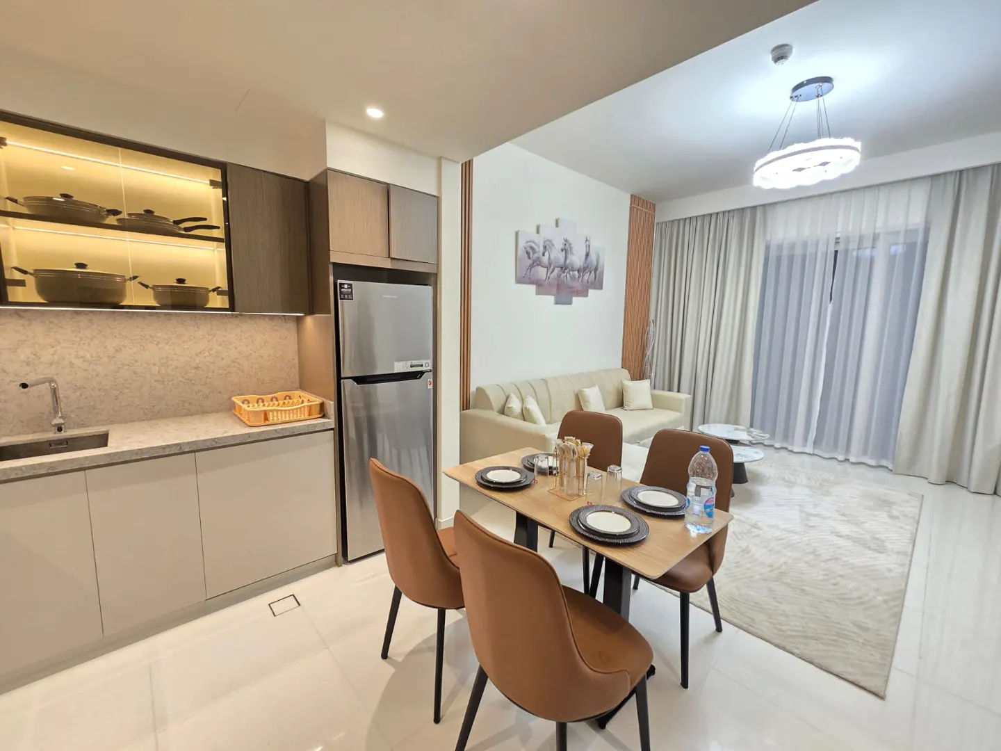A modern apartment interior with a kitchen, dining table, and living room. The kitchen features light cabinets, a stainless steel refrigerator, and a display cabinet. The dining table is set for four. The living room has a beige sofa and a large window.
