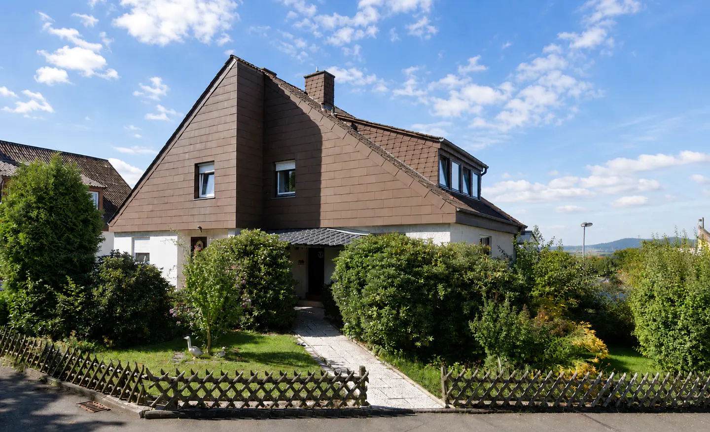 A two-story house with brown roof and white walls, surrounded by green bushes and a wooden fence.