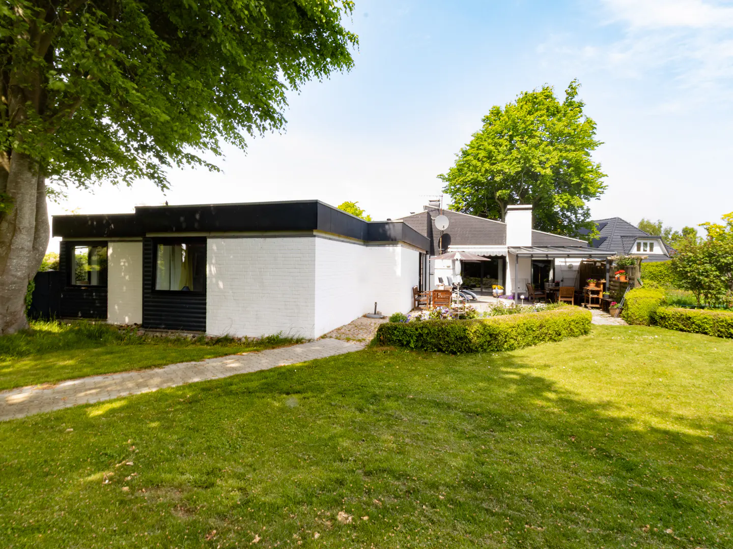 Modern white and black house with a paved path, lush green lawn, and a patio area surrounded by trees and hedges.