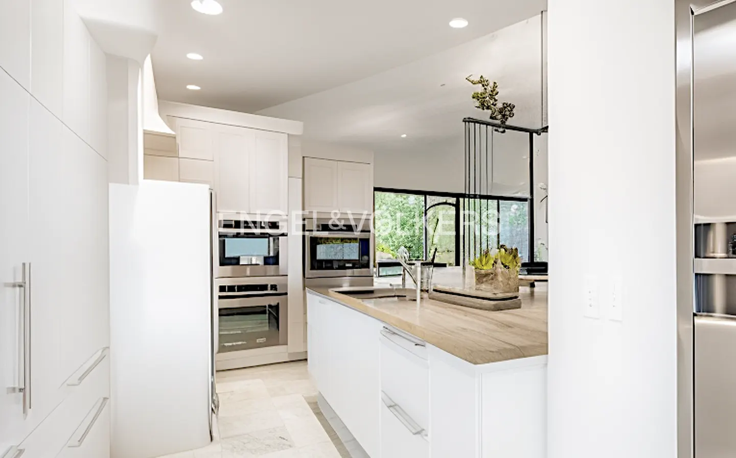 Bright, modern kitchen with white cabinets, stainless steel appliances, and a wood-topped island. Large windows offer natural light.