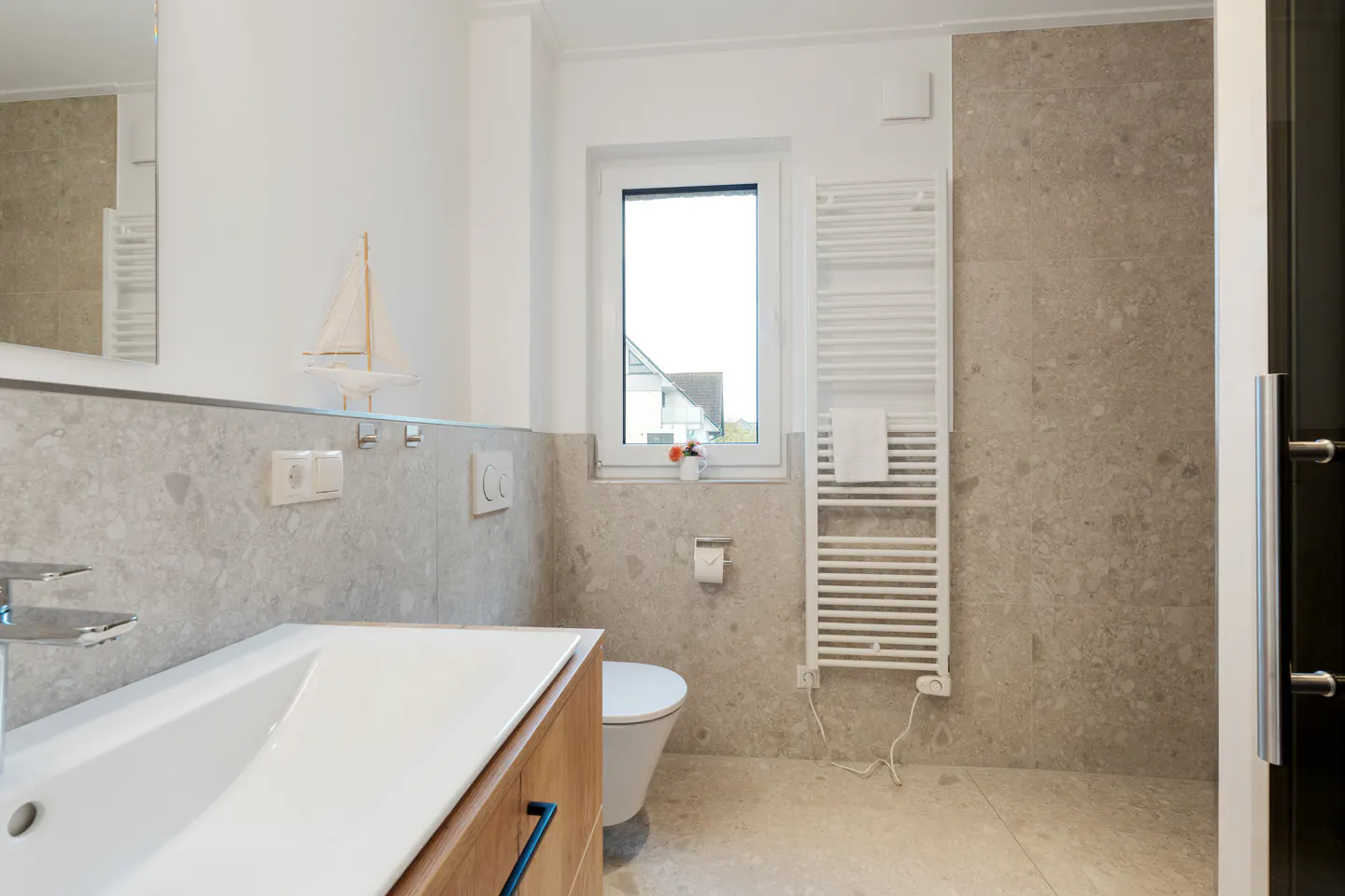 Bright bathroom with gray stone walls, white sink, wood cabinet, toilet, and towel warmer. A small sailboat sits on the counter.