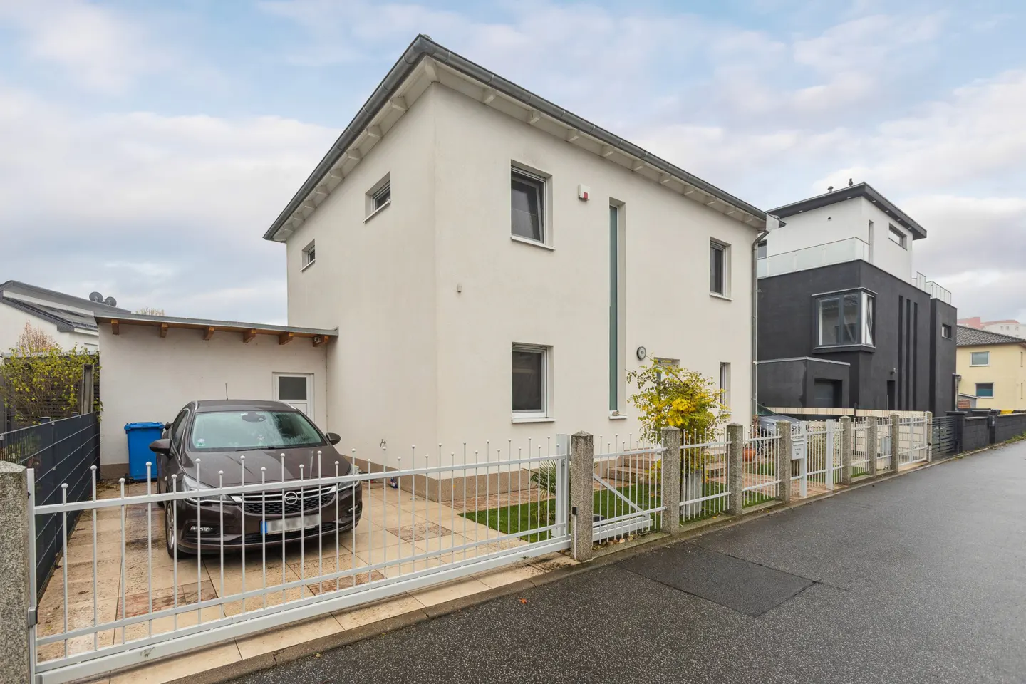 Exterior view of a modern two-story house with a white facade and a brown car parked in the driveway behind a white metal gate.
