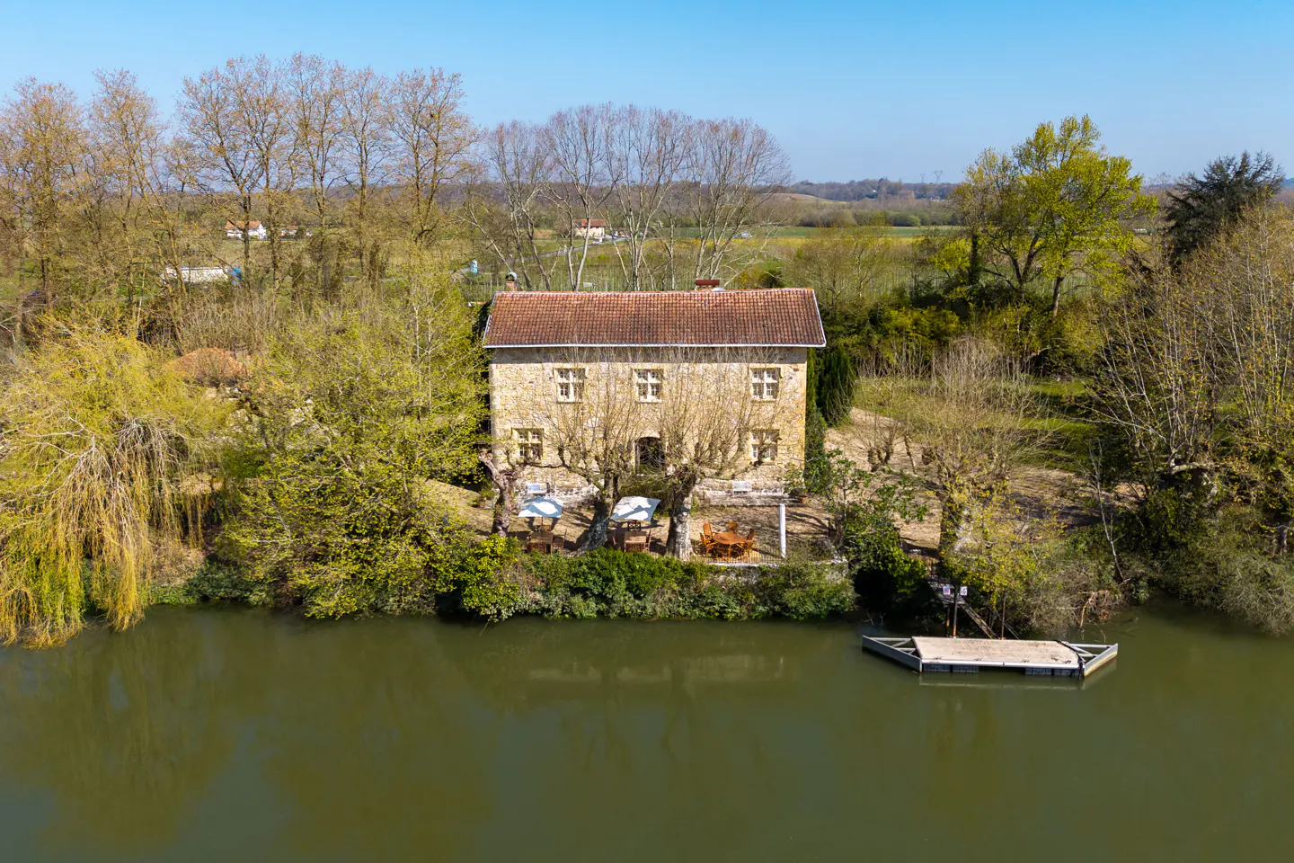 Two-story stone house with a red tile roof on a riverbank, surrounded by trees. A small dock floats on the water.