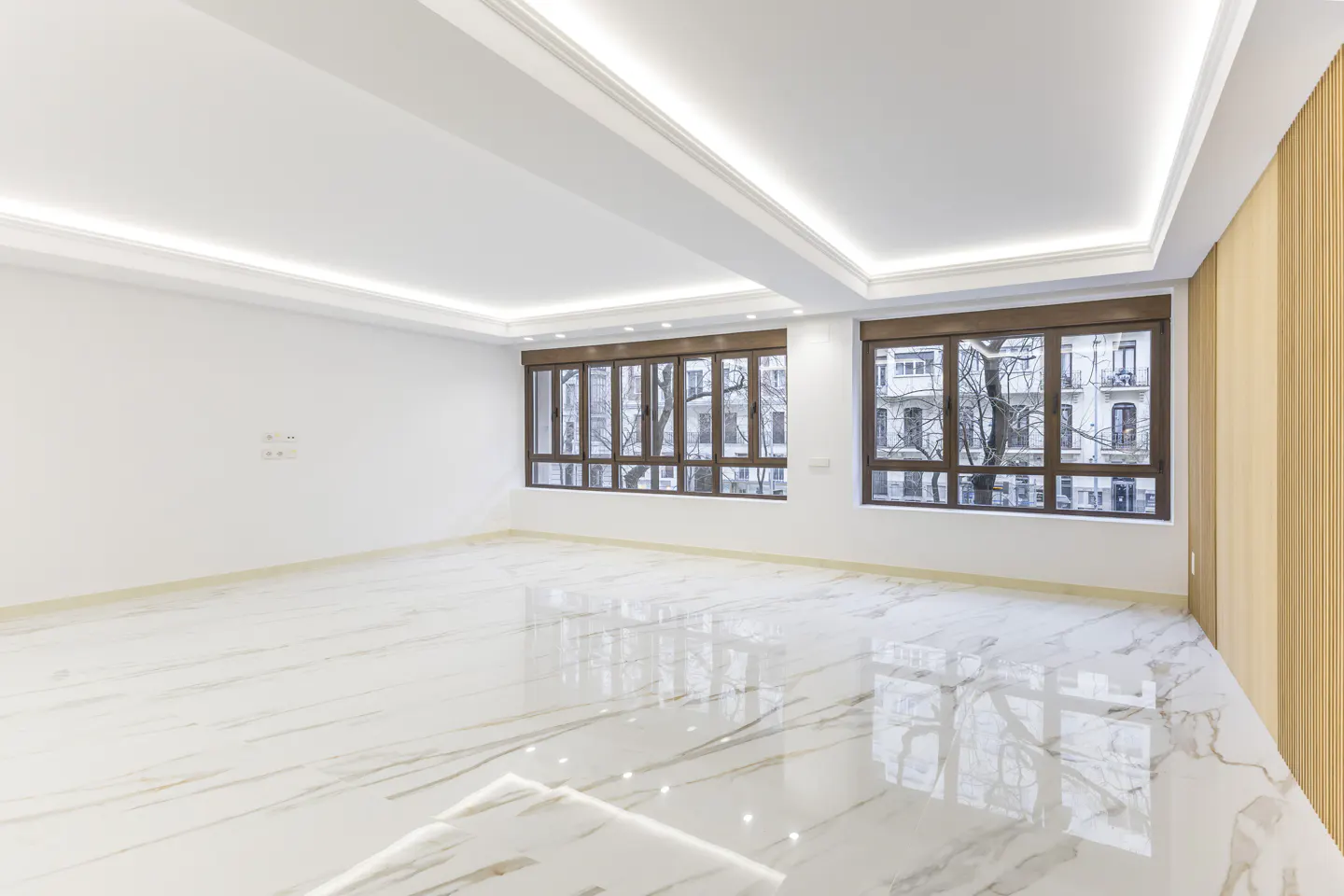 Bright, empty room with white walls, marble floors, and two large windows with brown frames. Outside, buildings and bare trees are visible.