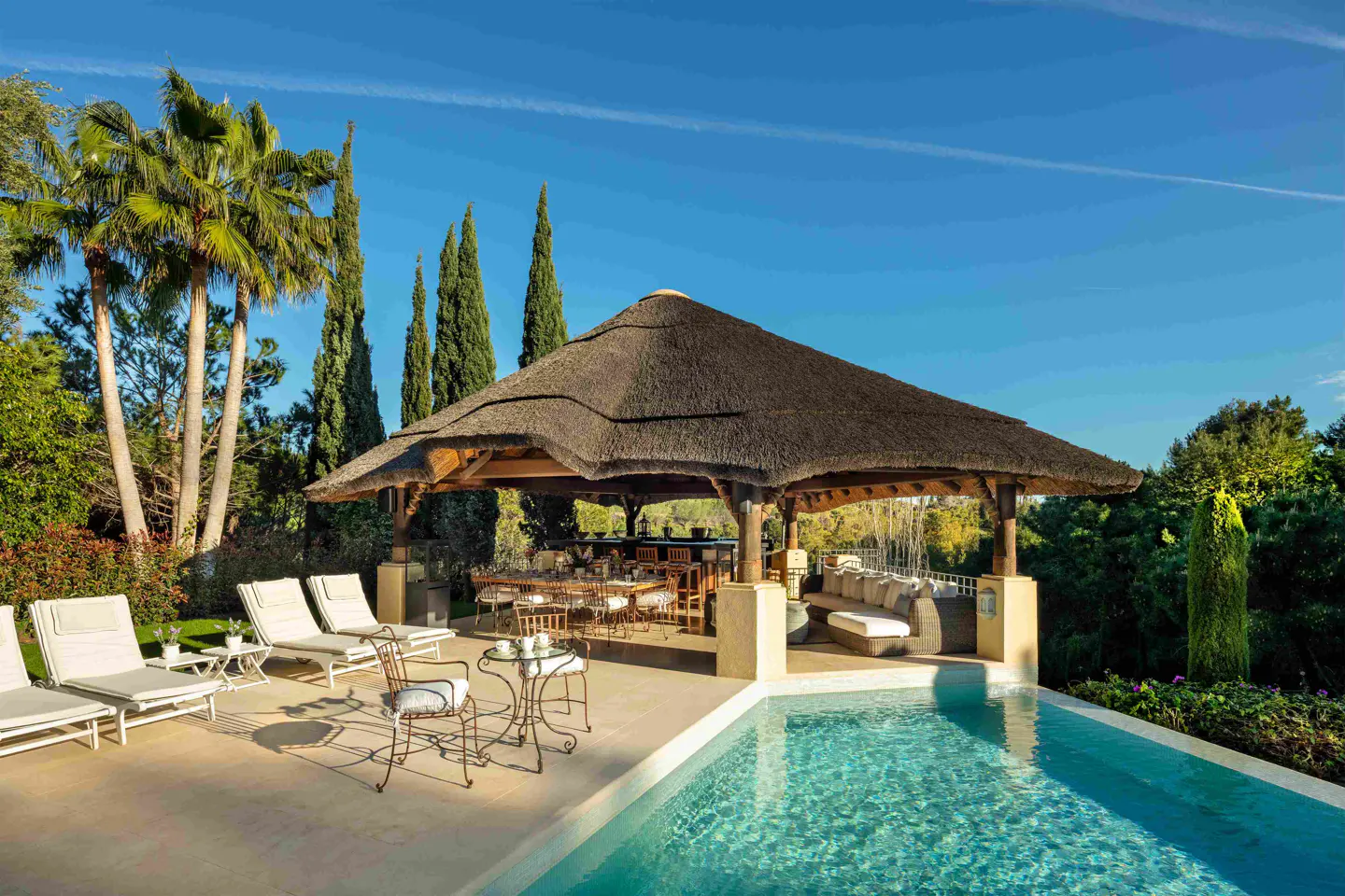 Luxury backyard with a pool, lounge chairs, and a thatched-roof gazebo with dining and seating areas. Palm trees and blue sky in the background.