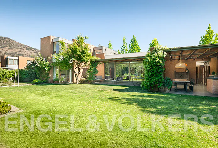 Exterior view of a modern, brown house with a large green lawn and patio furniture. Trees and a mountain are in the background.