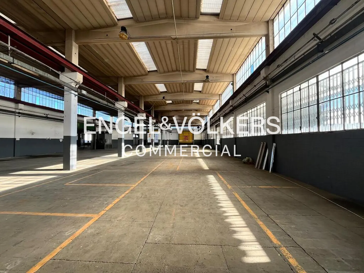 An empty warehouse with concrete floors, yellow lines, and large windows. The Engel & Völkers Commercial logo is superimposed on the image.