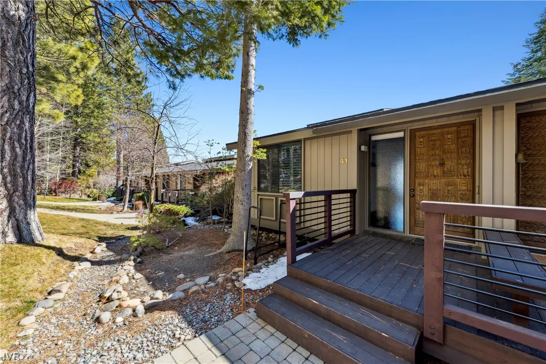 Exterior view of a tan condo with a wooden deck and steps, surrounded by trees and a rock-lined stream.