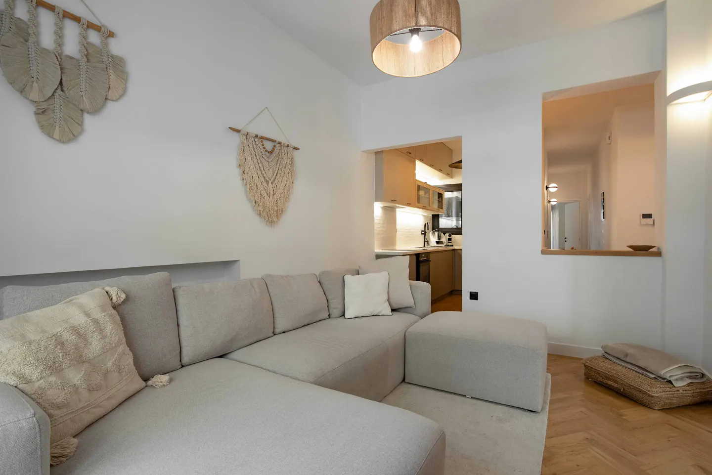 Living room with a beige sectional sofa, macrame wall art, and a view into the kitchen and hallway.