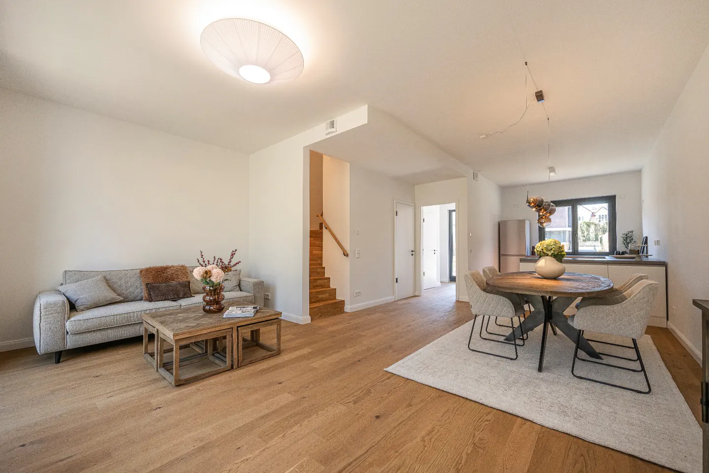 Bright, open-concept living space with wood floors, a gray sofa, and a round dining table with upholstered chairs. Stairs lead to the upper level.