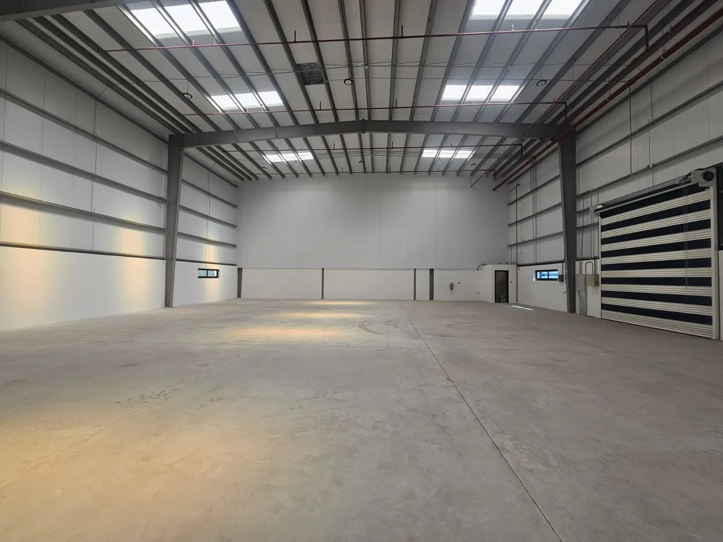 Empty warehouse interior with concrete floor, white walls, and a metal ceiling with skylights. A large, striped door is on the right.