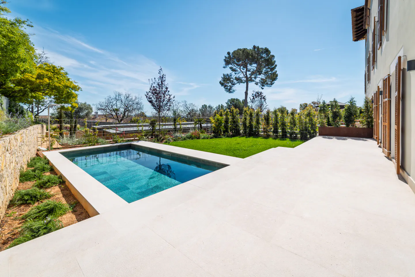 A rectangular pool with turquoise water is surrounded by a white stone patio and green lawn. Trees and a stone wall are in the background.