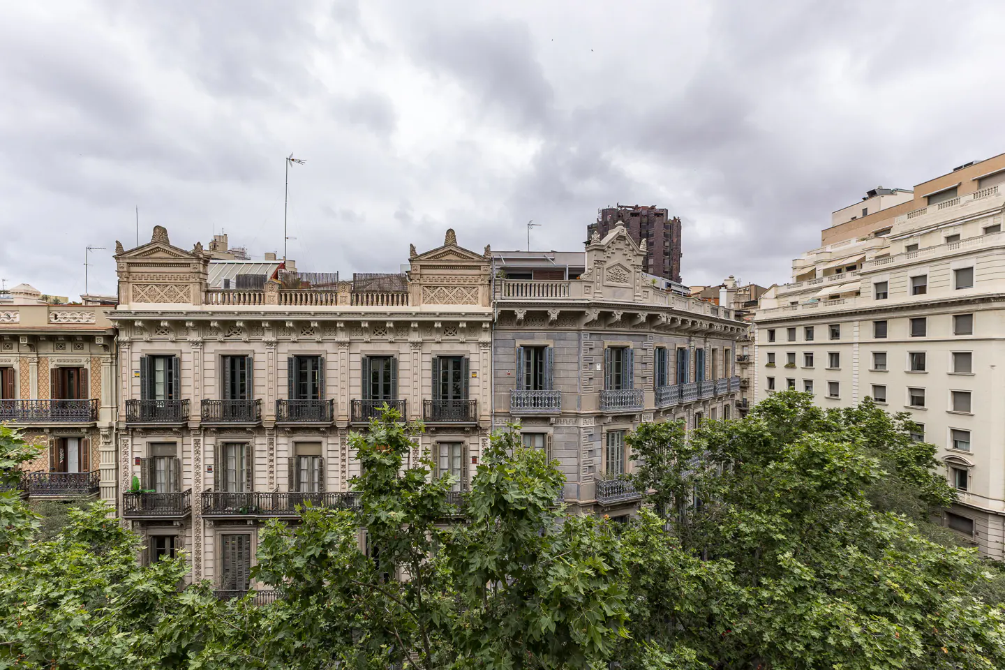 View of ornate, historic European apartment buildings with balconies and shutters, partially obscured by lush green trees.