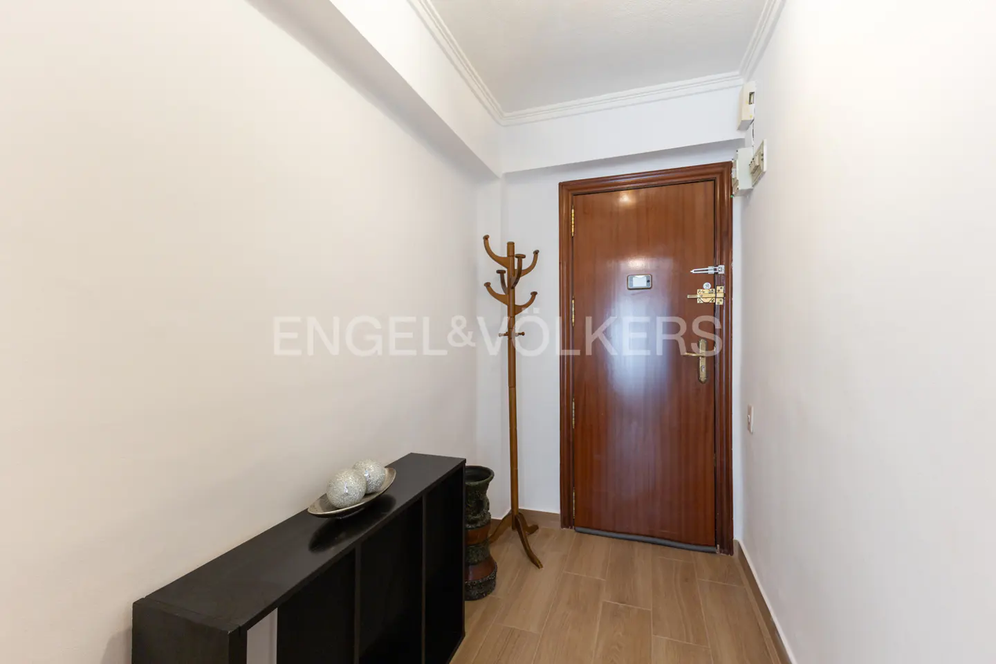 A bright hallway with white walls, a wooden door, a black console table with decorative spheres, and a wooden coat rack.
