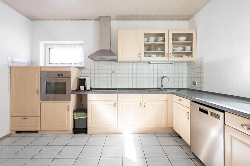 Bright kitchen with light wood cabinets, stainless steel appliances, and white tile backsplash. Dishes are visible in glass-front cabinets.