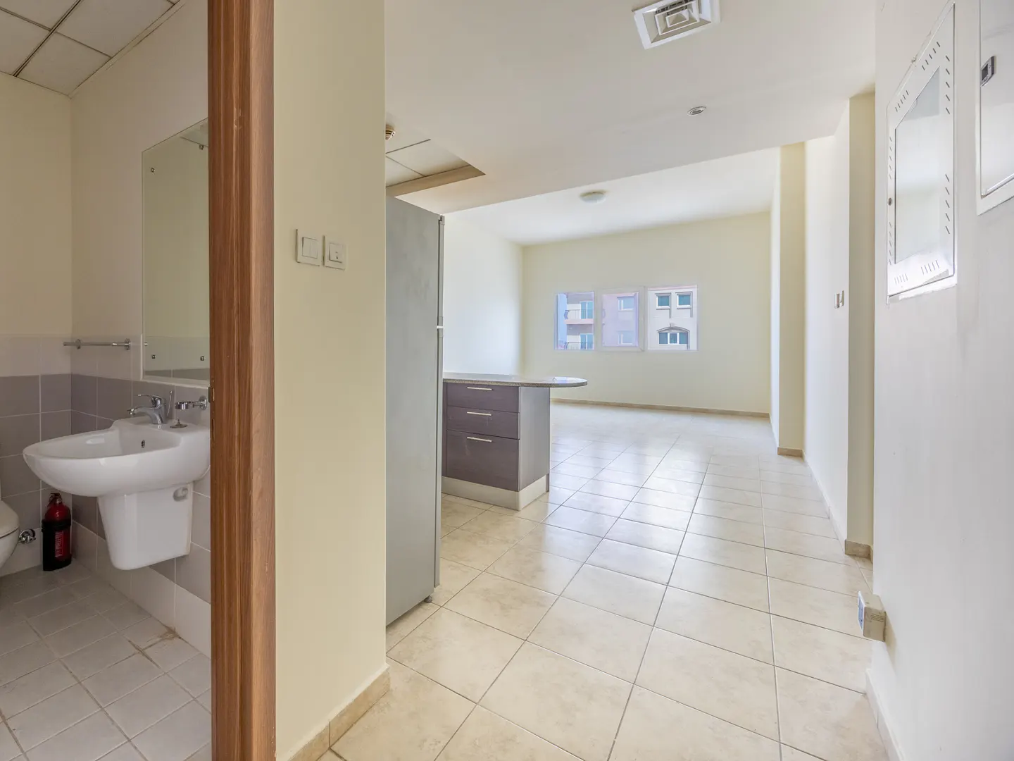 A bright apartment interior with beige tile floors, a bathroom with a white sink, and a kitchen island with brown cabinets.