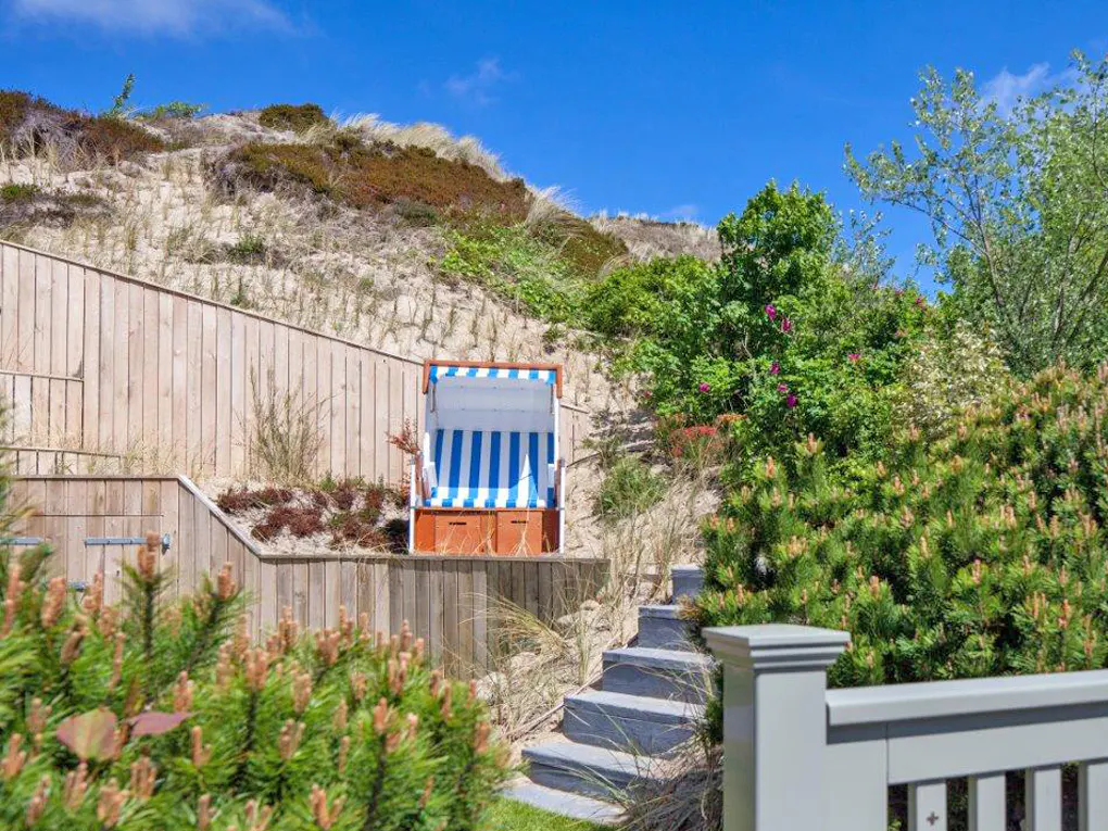 Outdoor scene with a blue and white striped beach chair on a wooden platform, surrounded by greenery and a sandy hill under a blue sky.