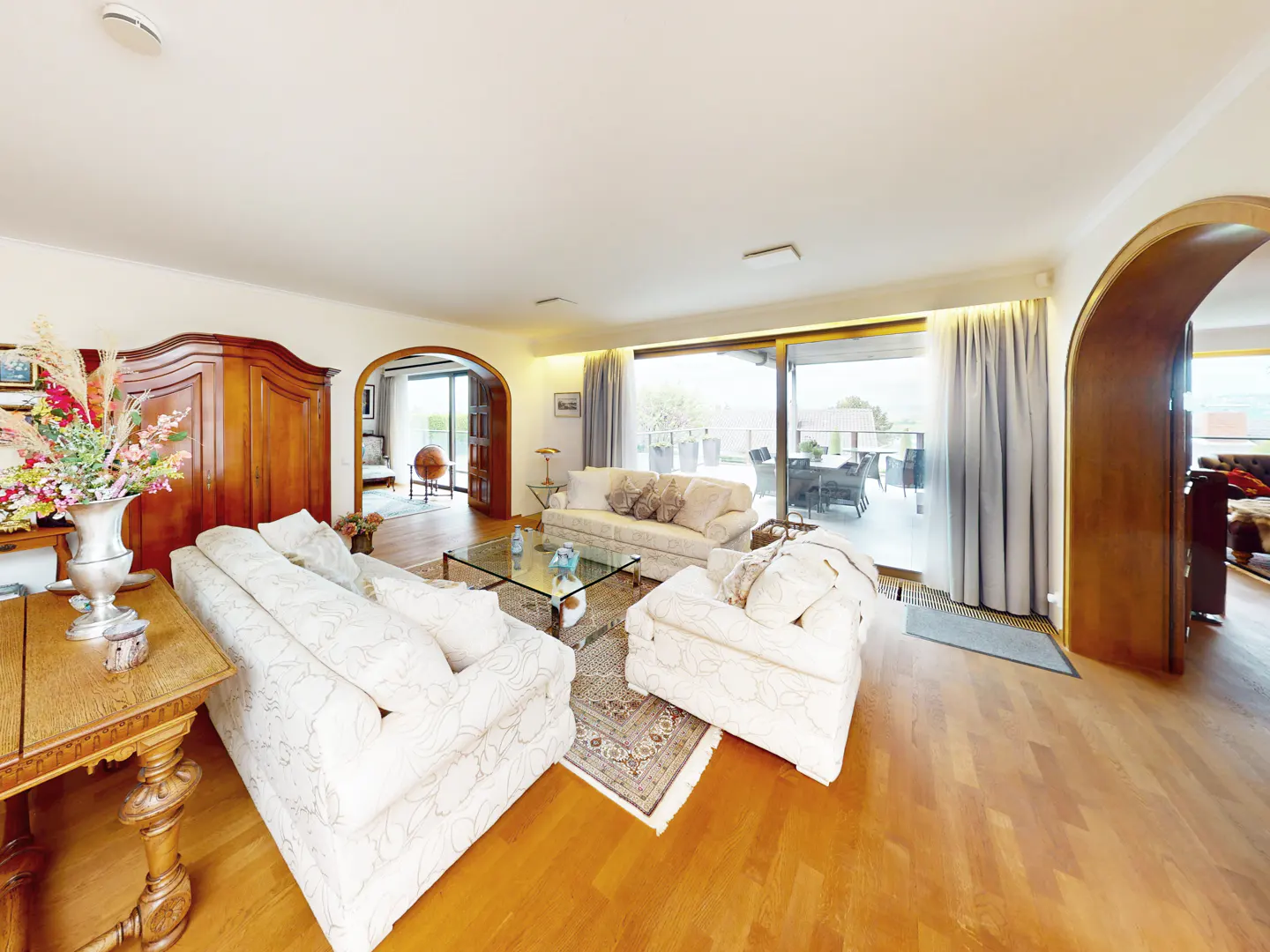 Bright living room with hardwood floors, white sofas, glass coffee table, and large windows leading to a patio.