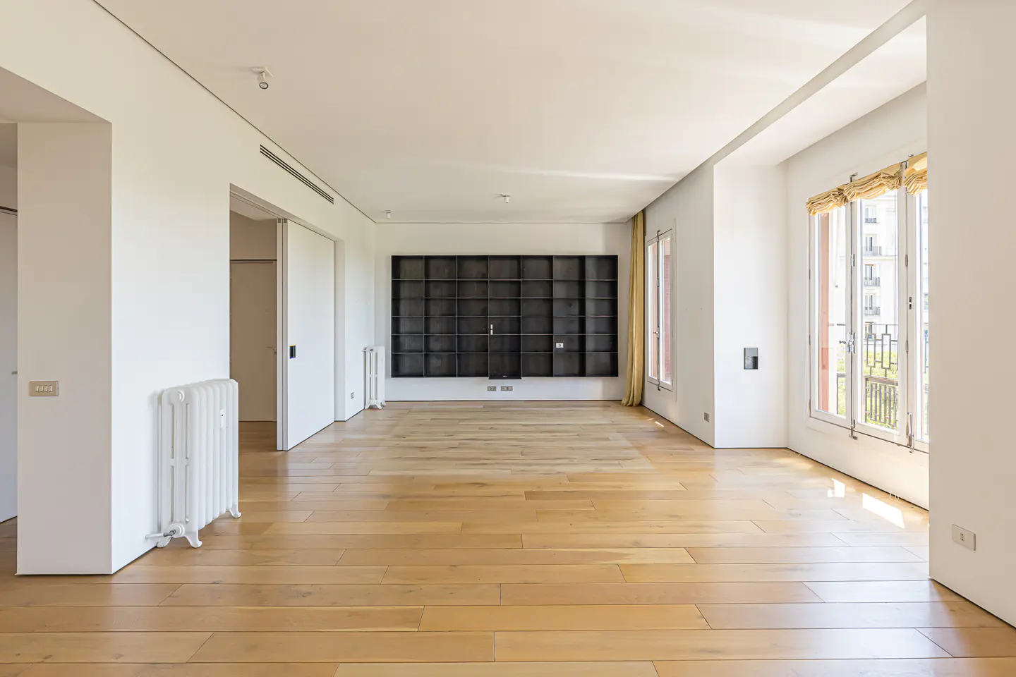 Bright, empty room with light wood floors, white walls, and a large, dark bookshelf. Windows with yellow curtains let in natural light.