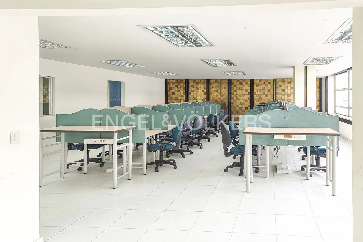 A bright office space with rows of desks and teal partitions, under fluorescent lighting.