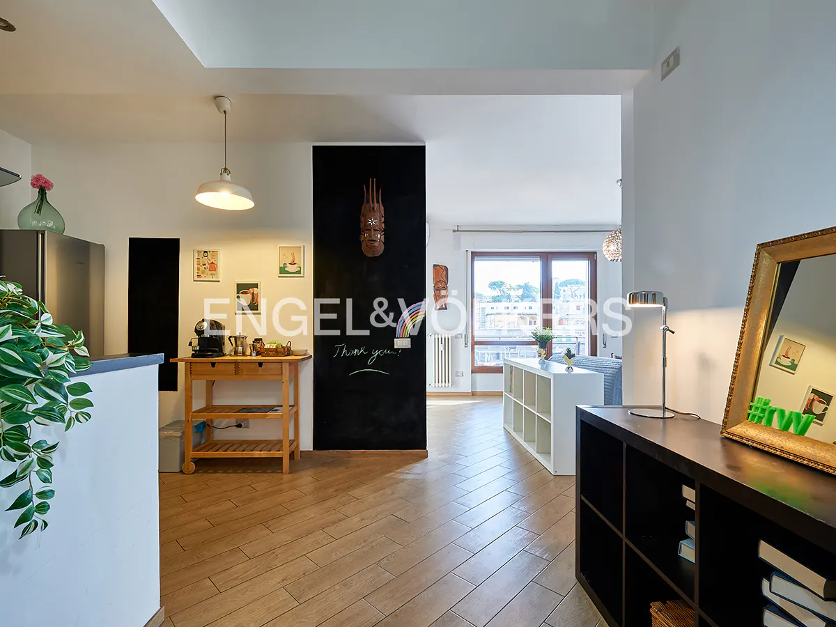 Interior view of a modern apartment with light wood floors, a black accent wall with "Engel & Völkers" logo, and a view into the living room.