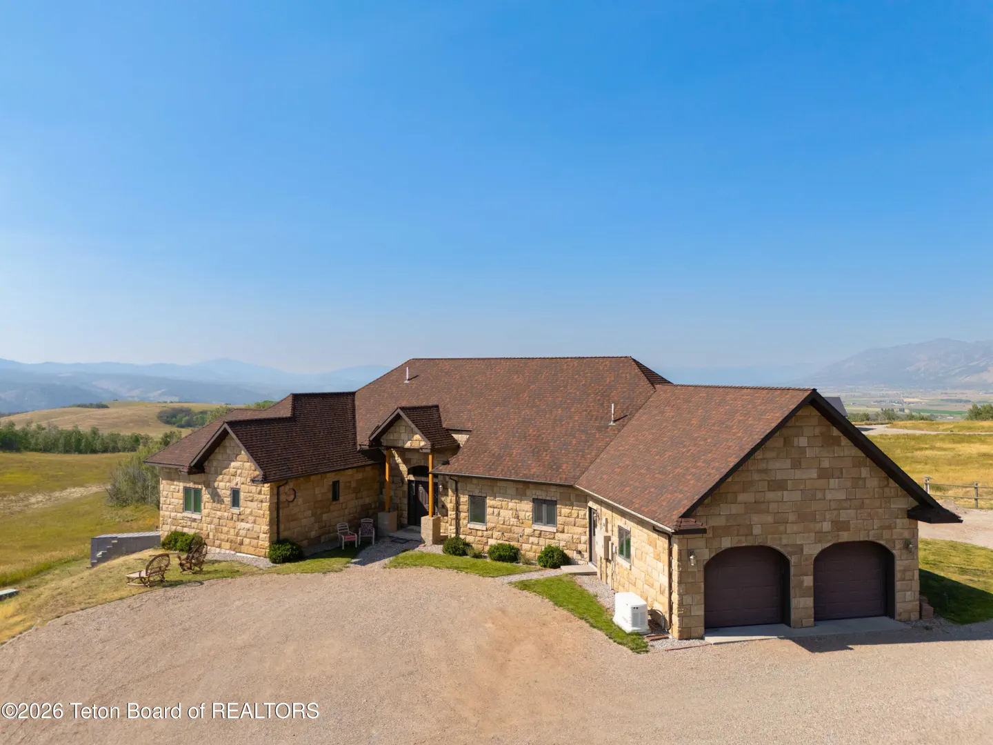 Aerial view of a tan stone house with a brown roof, a gravel driveway, and a mountain range in the background.