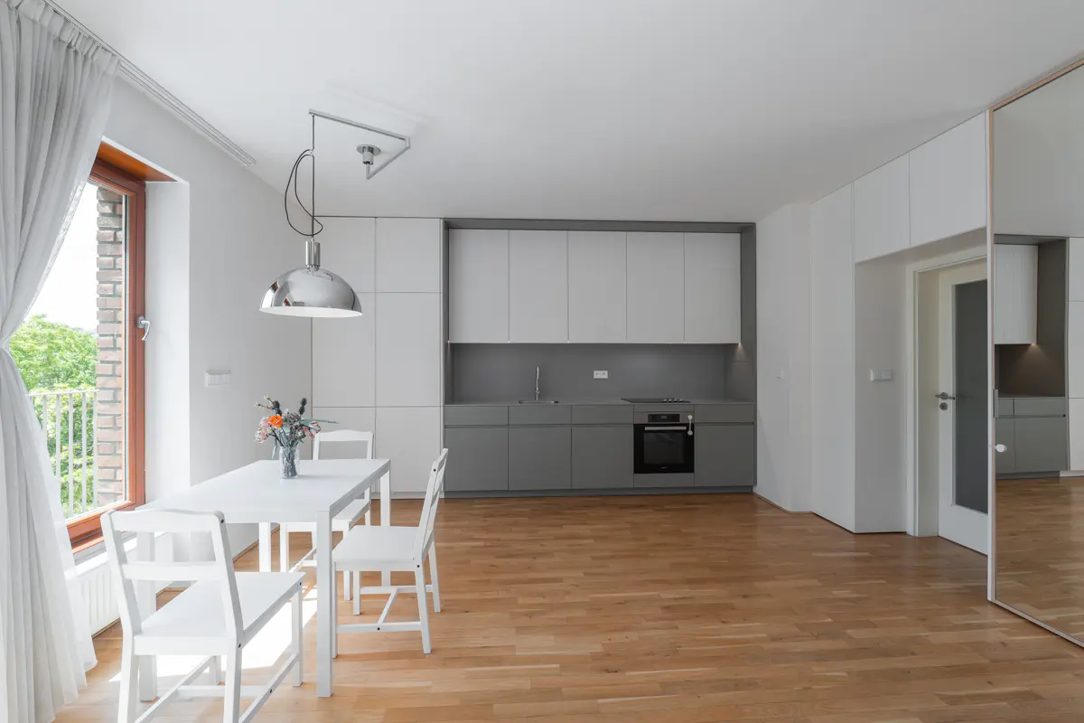 Bright, modern apartment with white walls, wood floors, and gray/white kitchen. A white table with chairs sits near a window with white curtains.