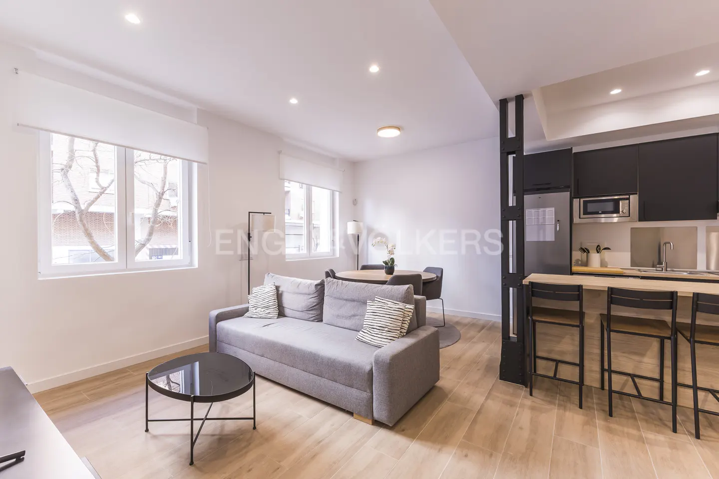 Modern open-plan living room with a grey sofa, round coffee table, dining area, and a kitchen with black cabinets.