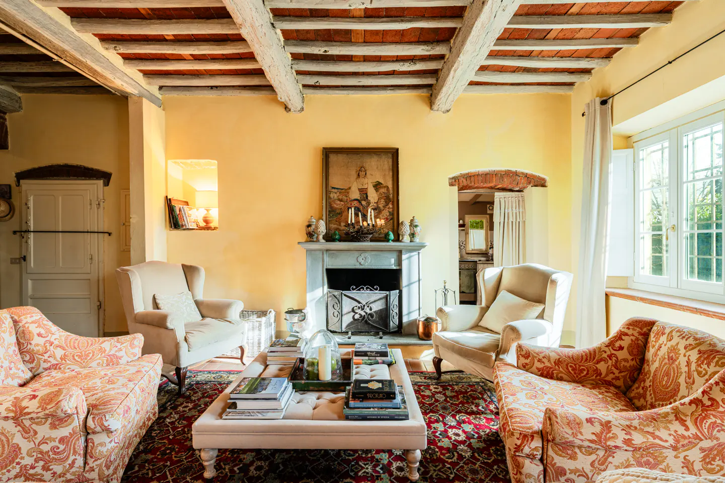 A living room with yellow walls, exposed beams, a fireplace, and floral armchairs.