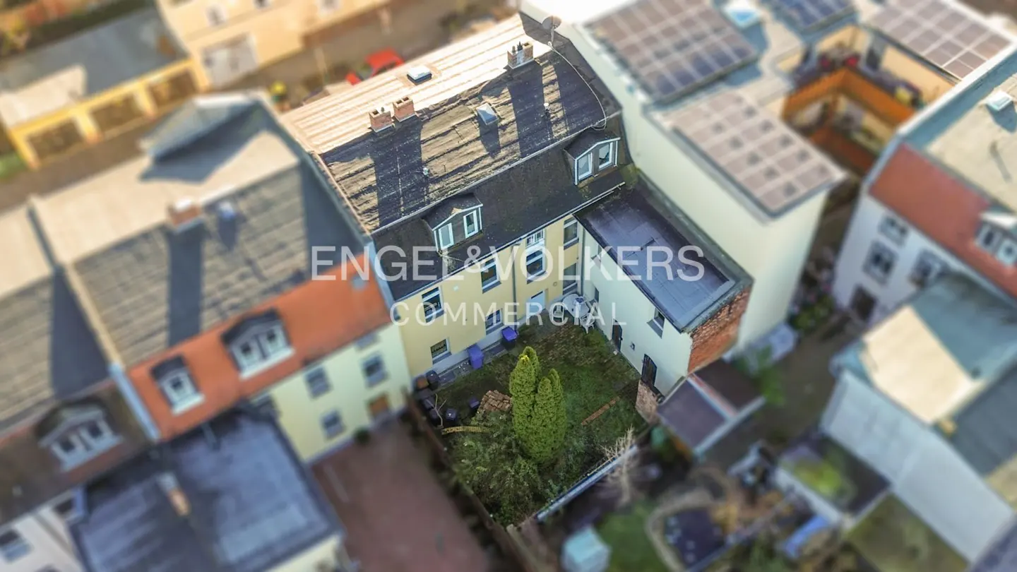 Aerial view of a yellow building with a black roof and a green backyard, surrounded by other buildings.