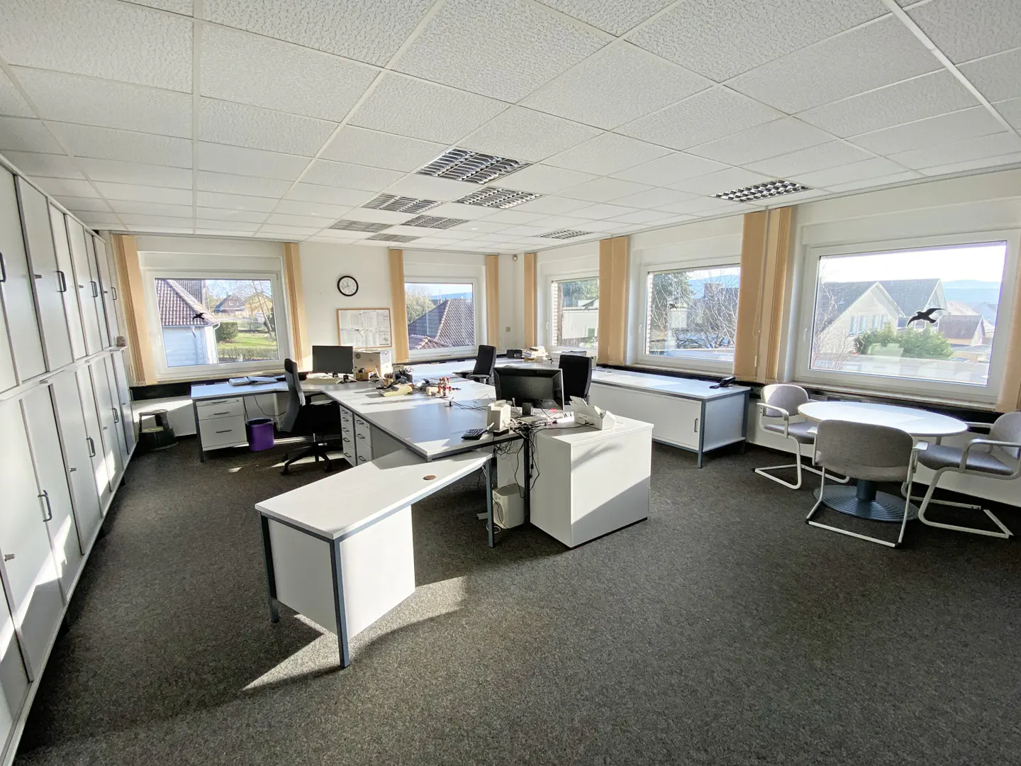 Bright office space with white desks, gray chairs, and gray carpet. Windows offer views of houses and trees. White lockers line the left wall.