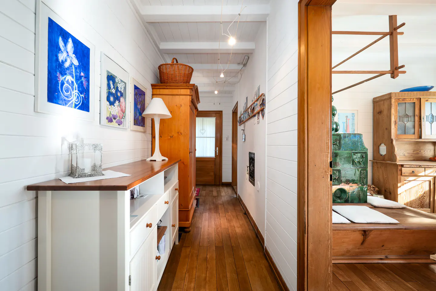 Hallway with white paneled walls, wood floors, and a white cabinet with a wood top. Blue floral art hangs above. A wood stove is visible in the next room.