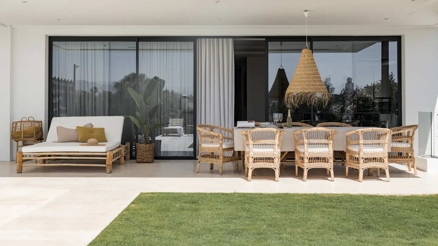 Outdoor patio with a long table and wicker chairs under a straw pendant light. A daybed sits to the left of the sliding glass doors.