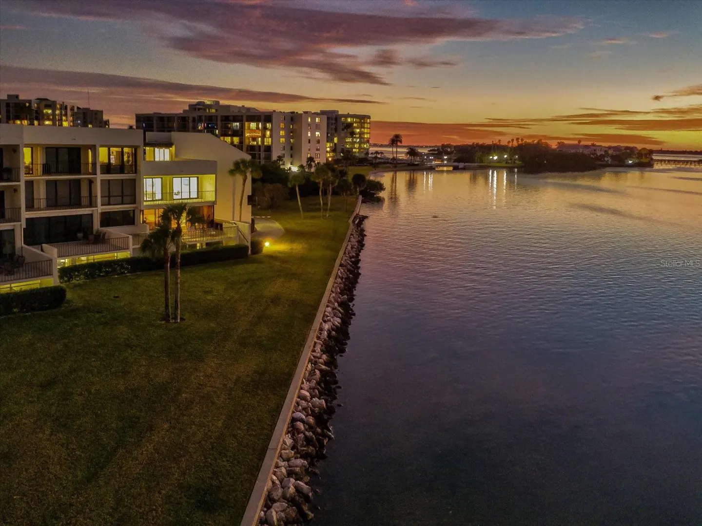Waterfront condos at sunset. Buildings are white with lit windows. Green lawn and palm trees line the water's edge. Sky is orange and purple.