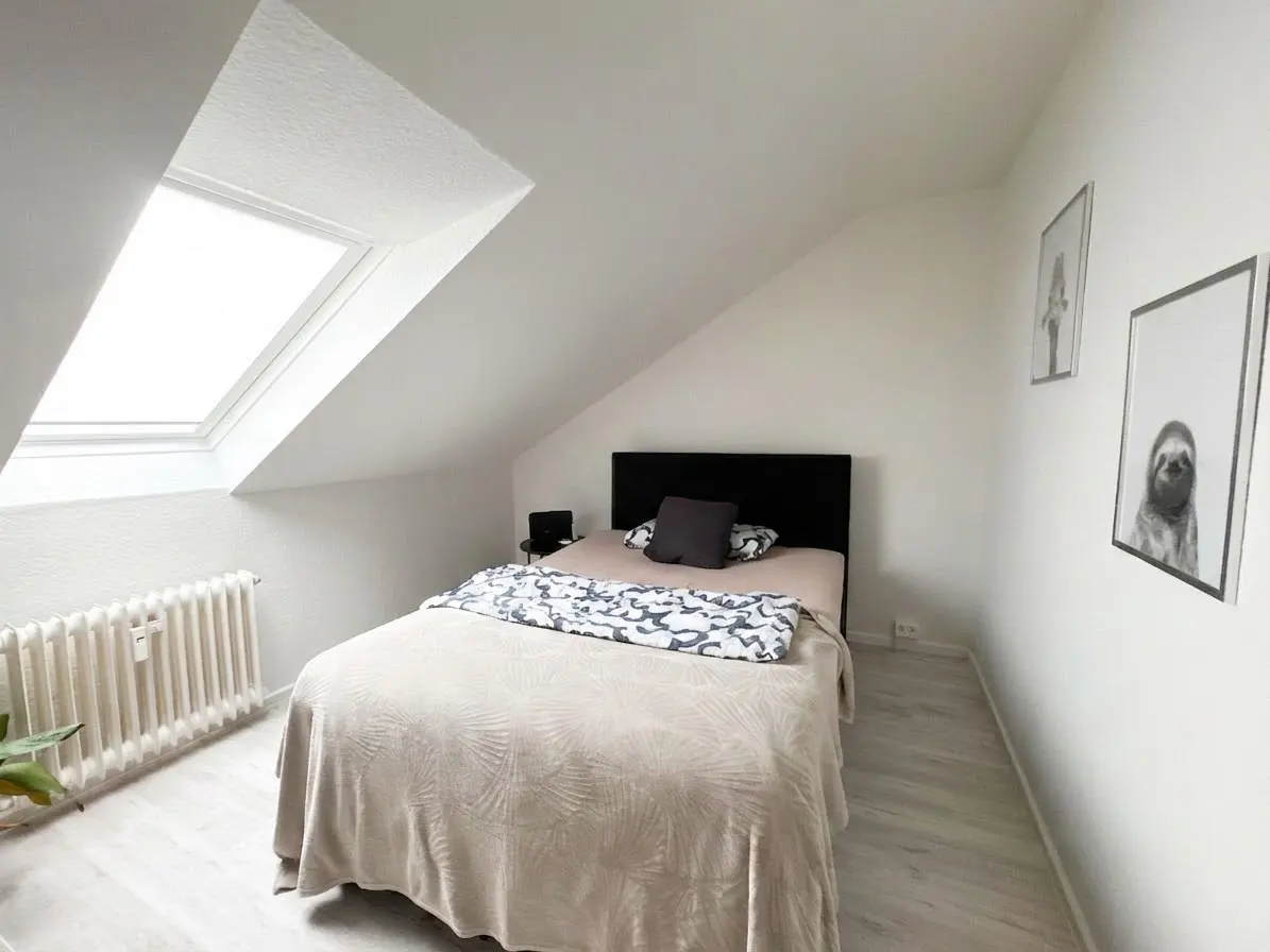 Attic bedroom with white walls, a skylight, and a bed with a black headboard. Two framed pictures hang on the wall.