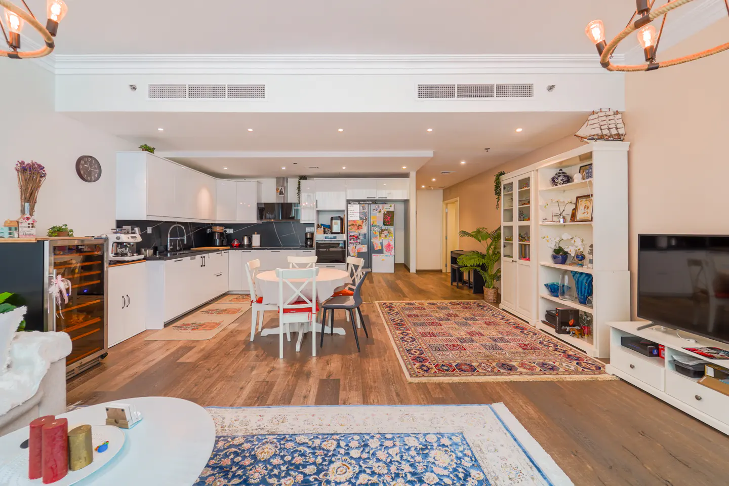 Open-concept living space with white kitchen, dining table, and living room. Hardwood floors and colorful rugs add warmth.