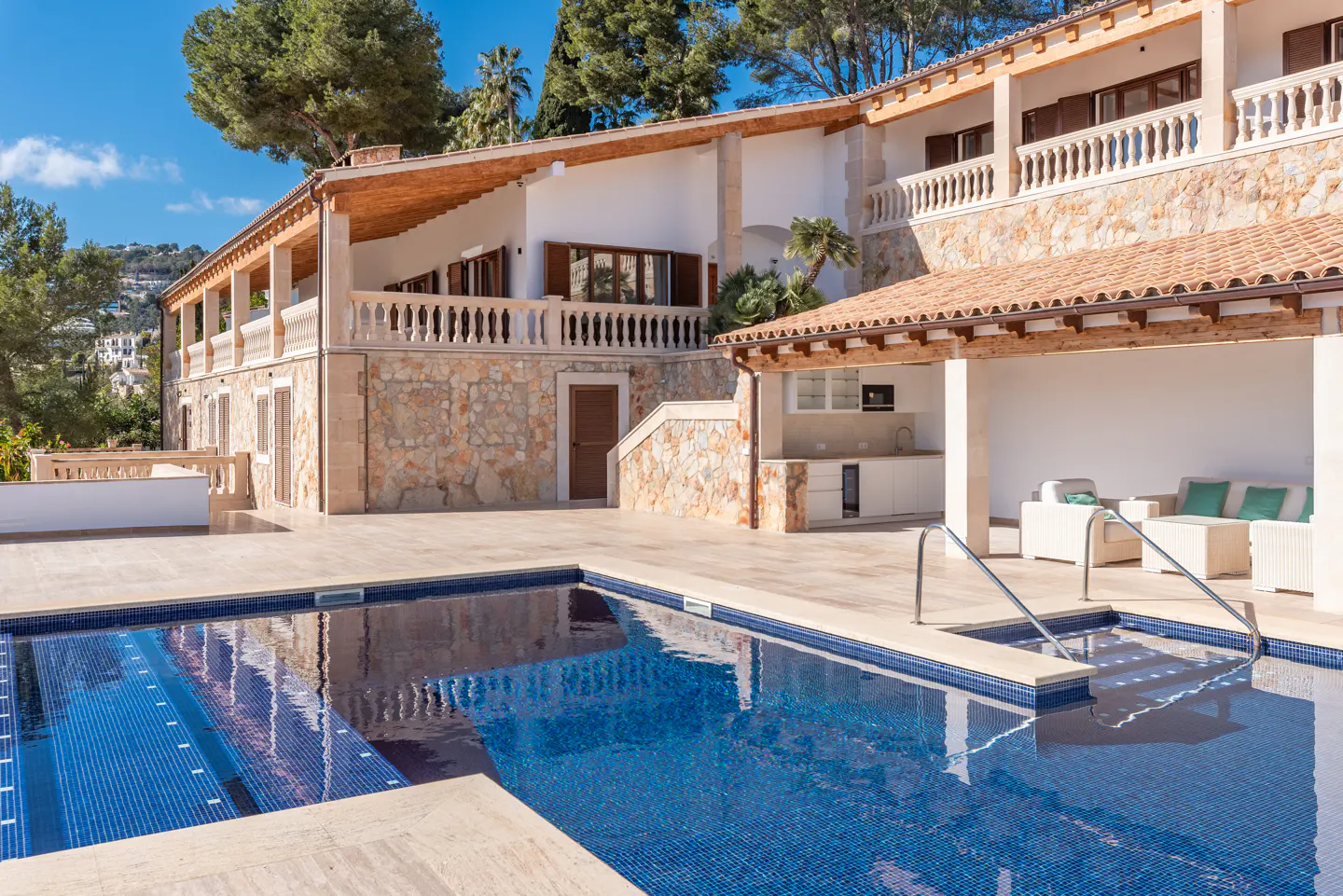 Exterior view of a stone house with a blue tiled pool, outdoor kitchen, and white patio furniture.