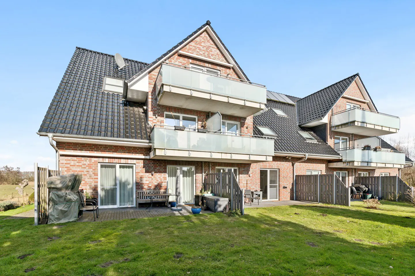 Brick apartment building with black roof, balconies, and green lawn. Patio furniture is visible on the ground floor.