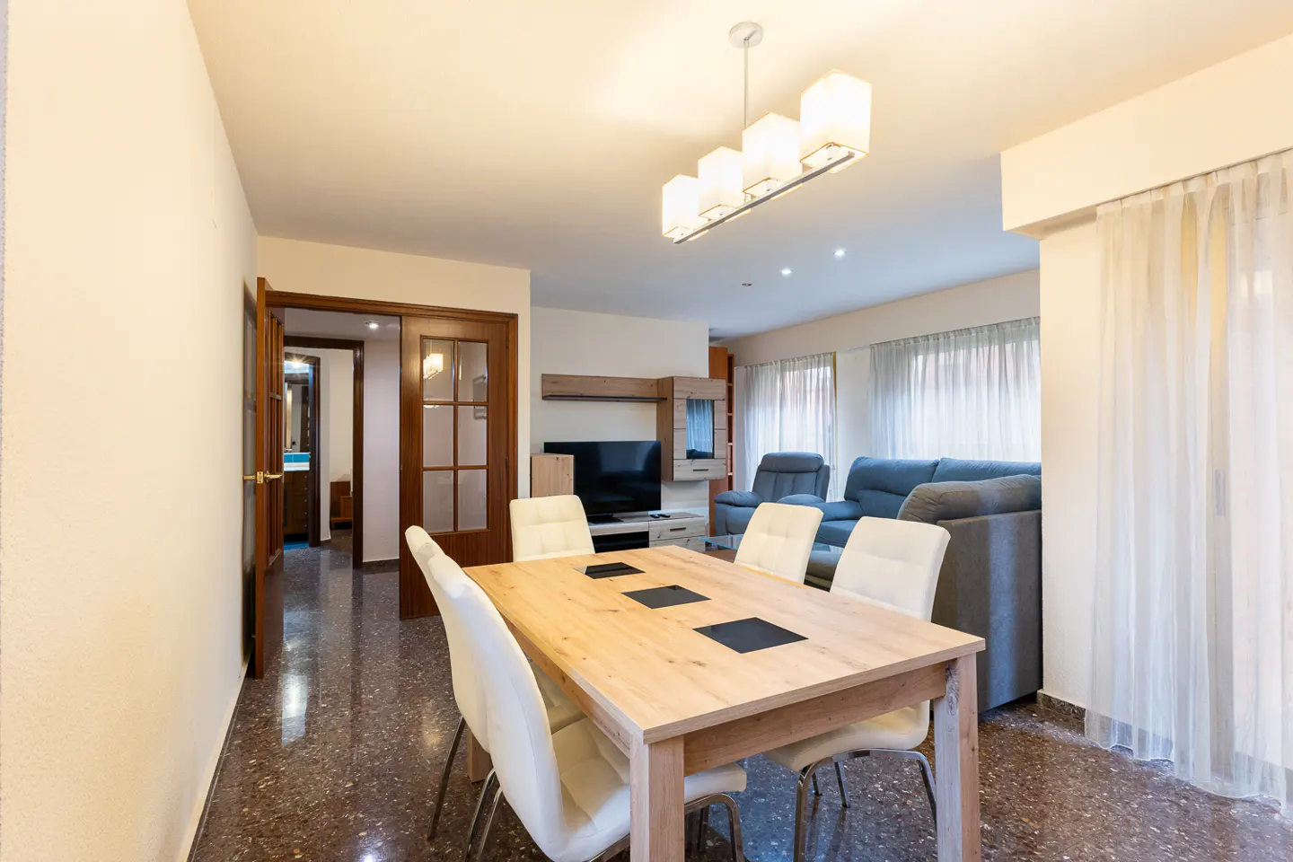 Bright dining area with a light wood table, white chairs, and a modern light fixture. Living room with blue sofas and a TV in the background.