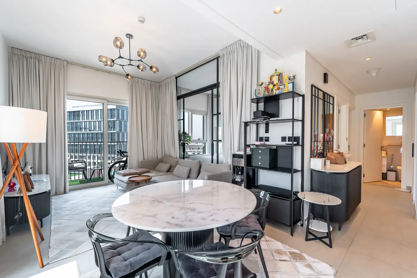 Modern dining area with a round marble table, transparent chairs, a sofa, and a balcony view in a bright, open-plan apartment.