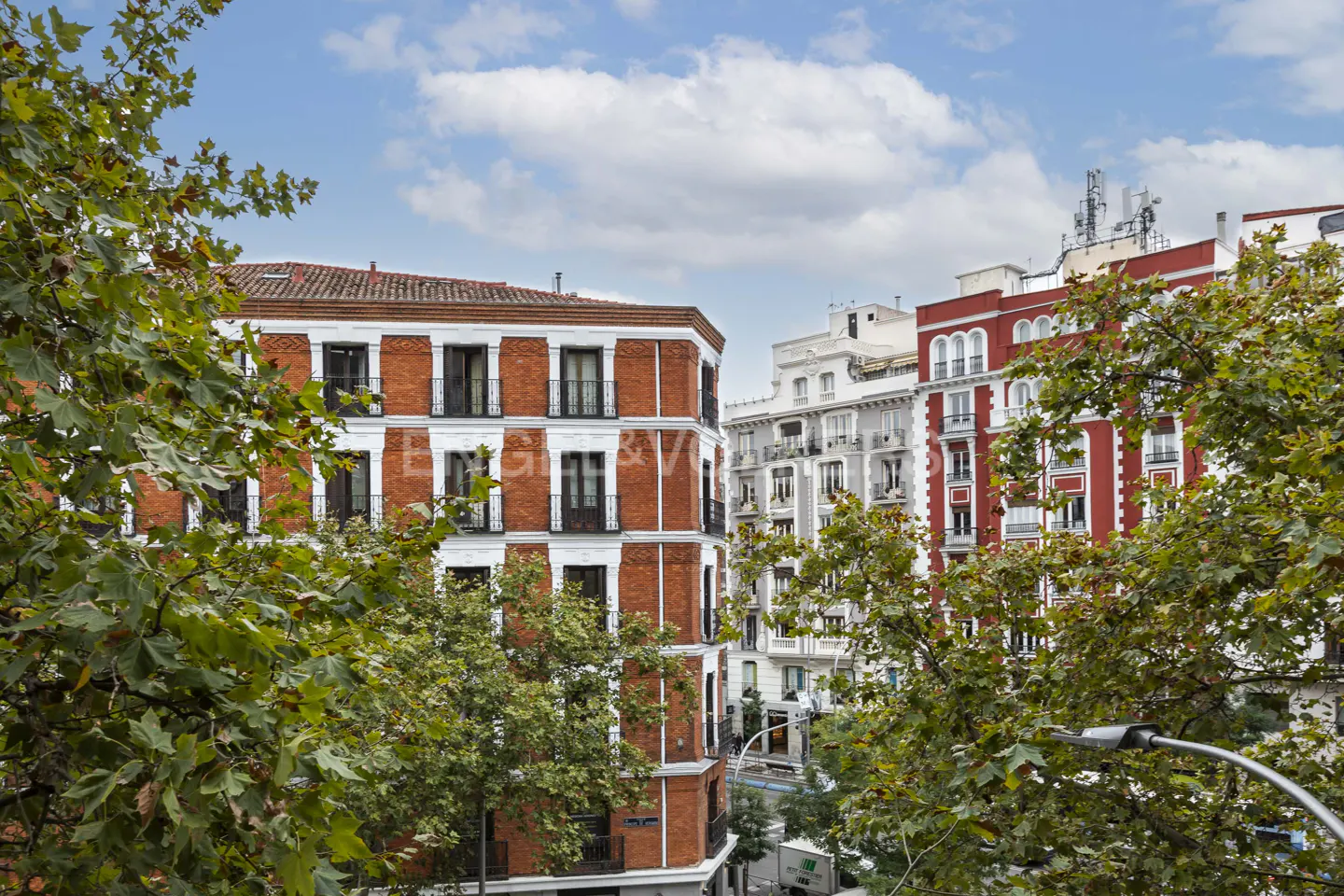 View of red brick buildings with black balconies, framed by green trees under a blue sky with clouds.