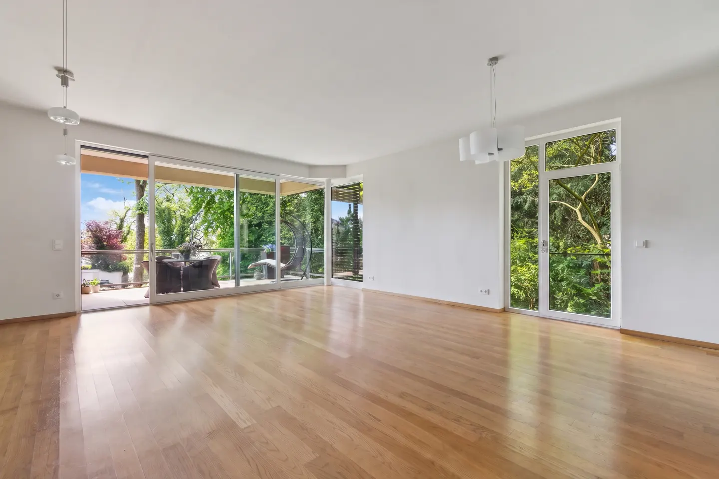 Bright, empty room with hardwood floors, white walls, and large windows. A patio with furniture is visible through the sliding glass doors.