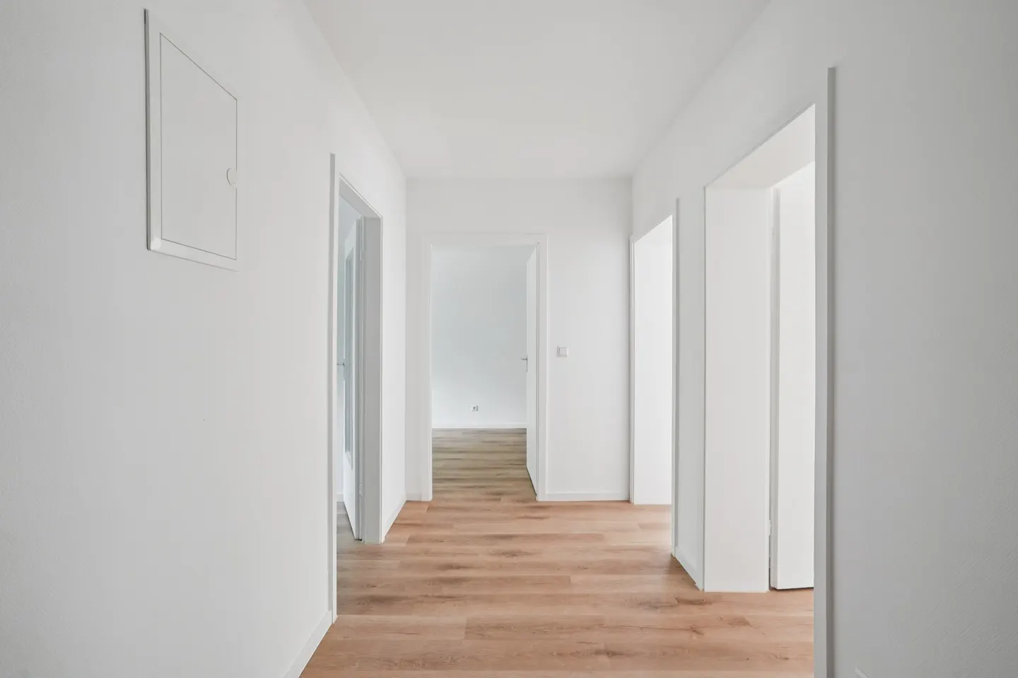 A bright, minimalist hallway with white walls and light wood flooring, featuring multiple doorways leading to other rooms.
