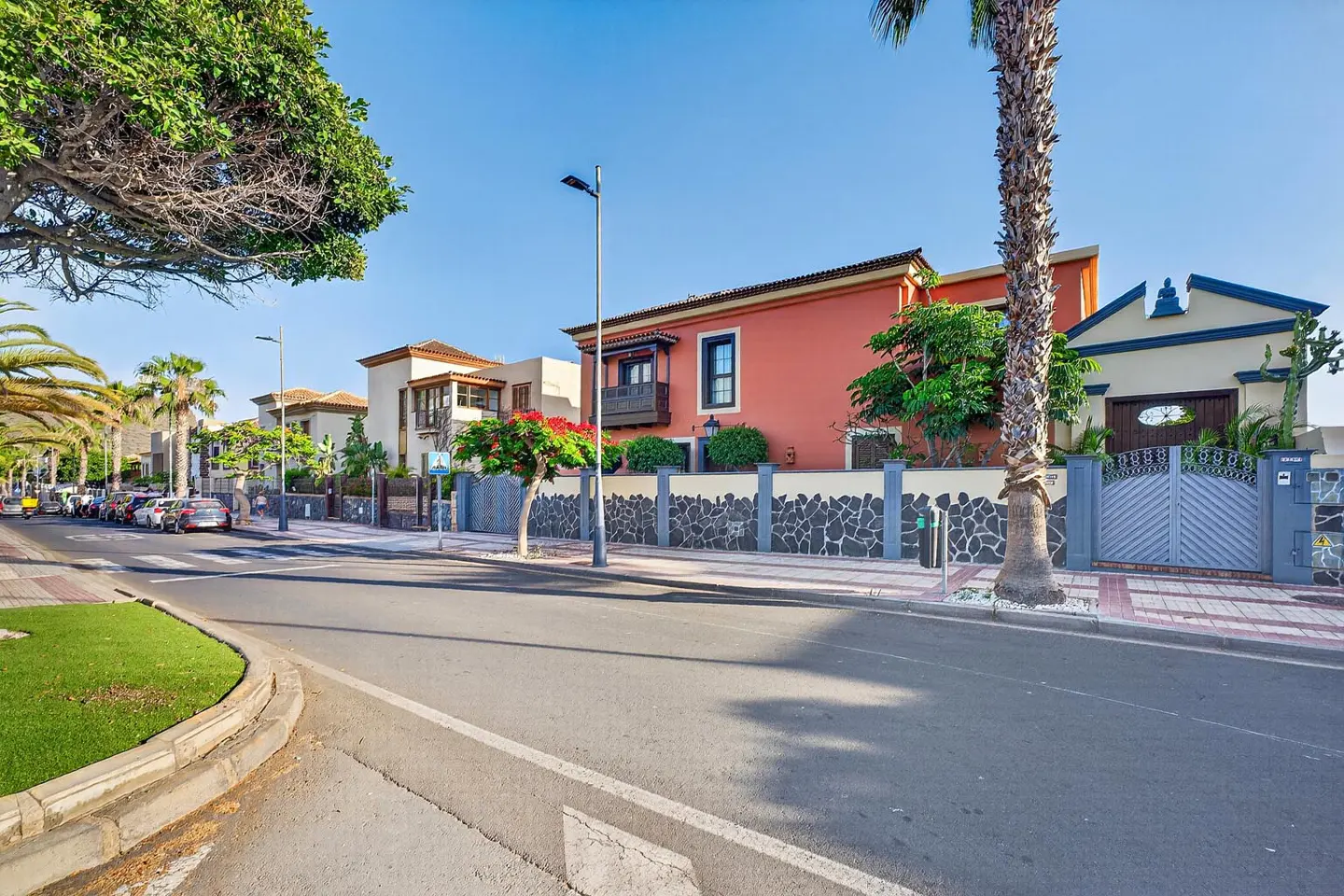 A sunny street view of a vibrant orange villa with a stone wall, palm trees, and a clear blue sky.